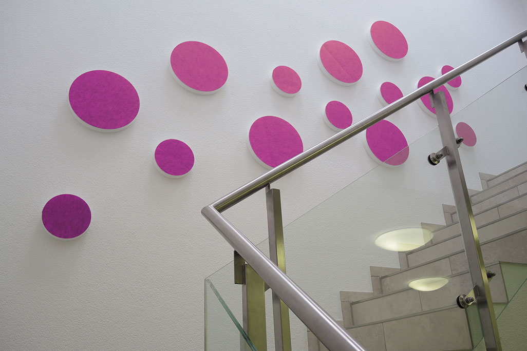 soni ELEMENT CIRCLE 14 Schallabsorber in Form eines Kreises (soni ELEMENT CIRCLE) in der Farbe Pink auf einer weißen Wand in einem modernen Treppenhaus angebracht.